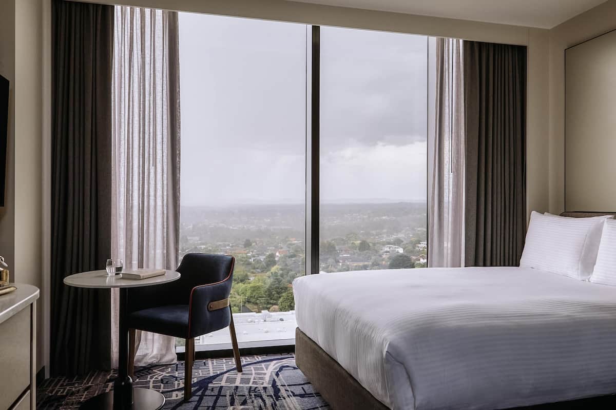 A hotel room with a large bed, a small table with a chair, and a view of a cityscape through large windows.