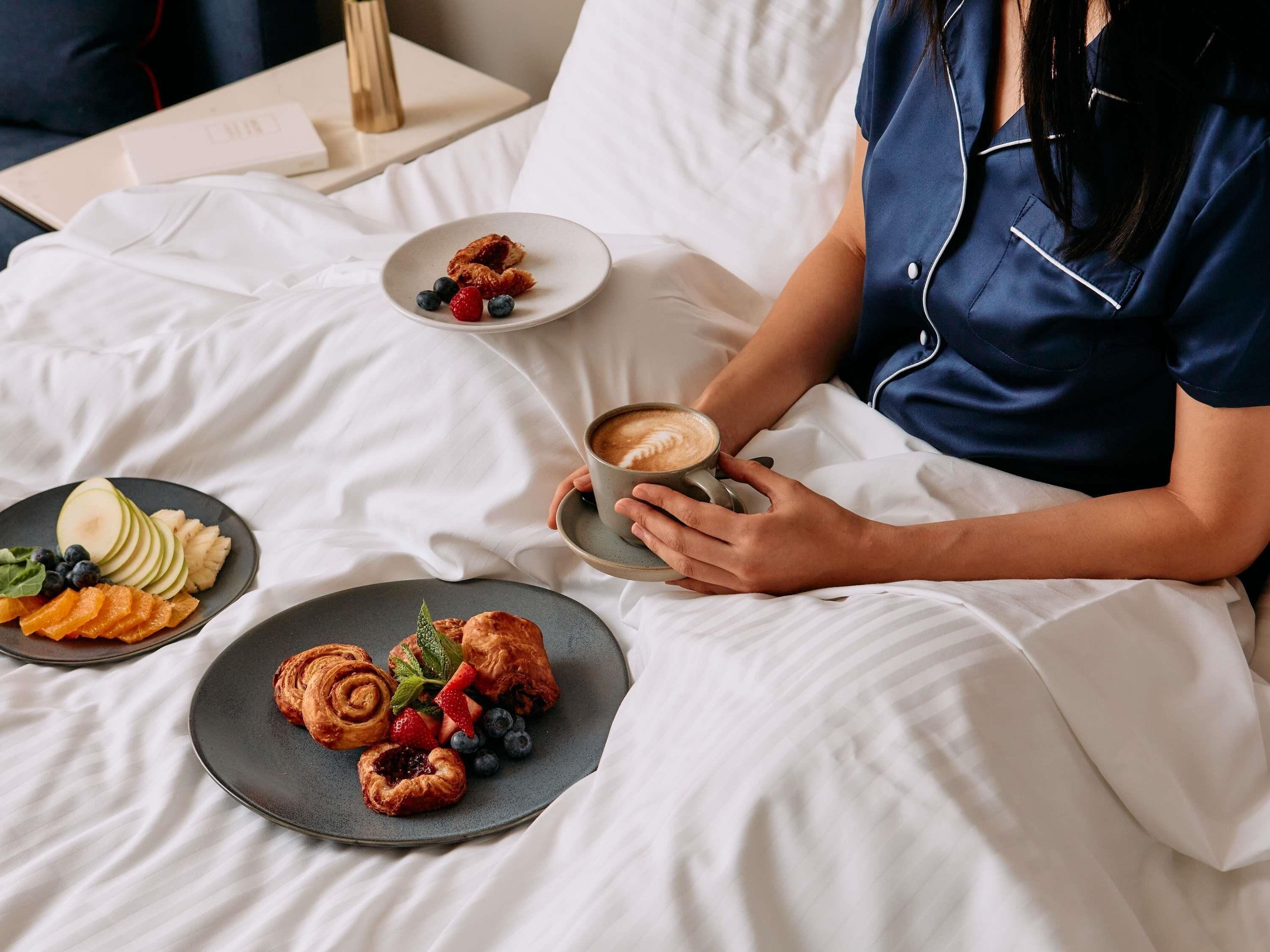 Eine Person in einem blauen Bademantel sitzt auf einem Bett mit weißer Bettwäsche und hält eine Tasse Kaffee. Auf dem Bett stehen Teller mit Gebäck und Obst.