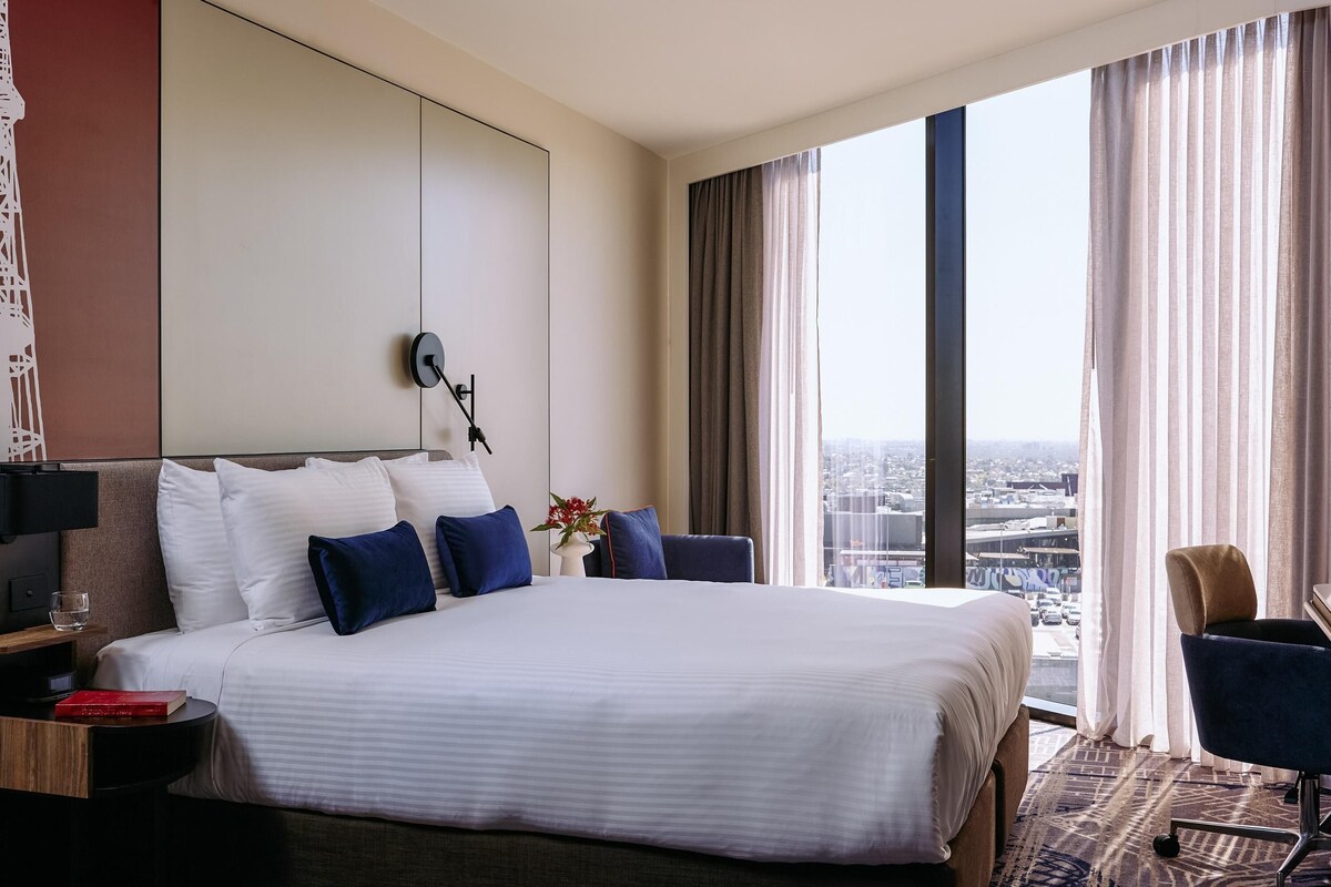A hotel room with a large bed, a desk, a chair, and a view of the city through large windows.
