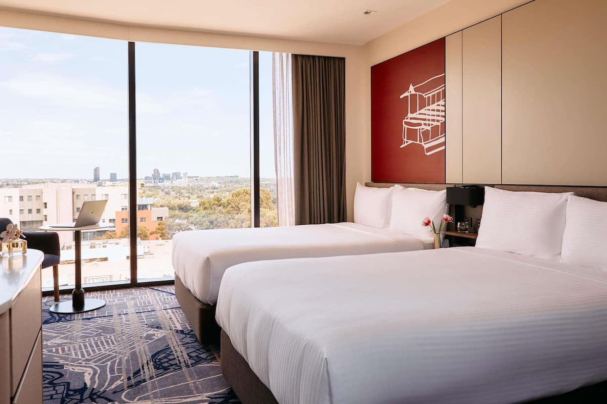 A hotel room with two beds, a desk with a laptop, a large window with a city view, and a red accent wall.