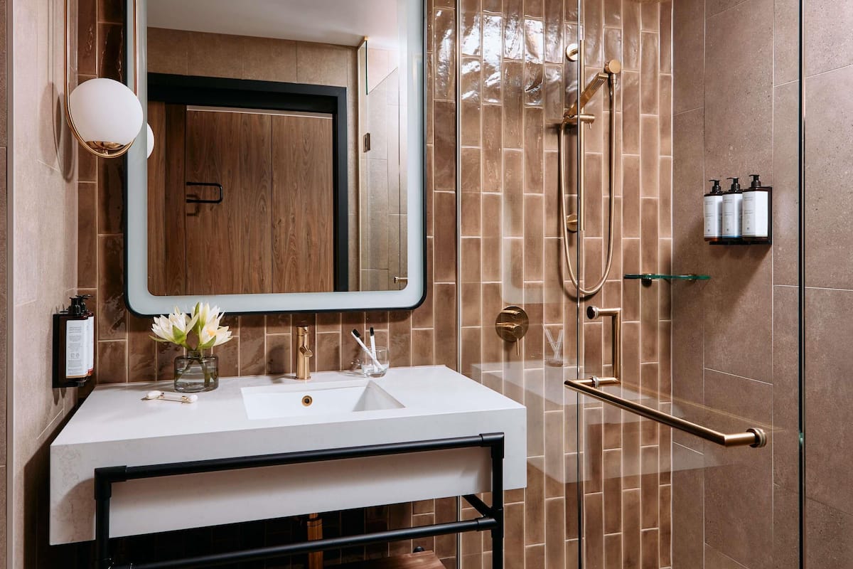 A modern bathroom with a large mirror, a sink, a shower with a handheld showerhead, and a glass shelf with toiletries.