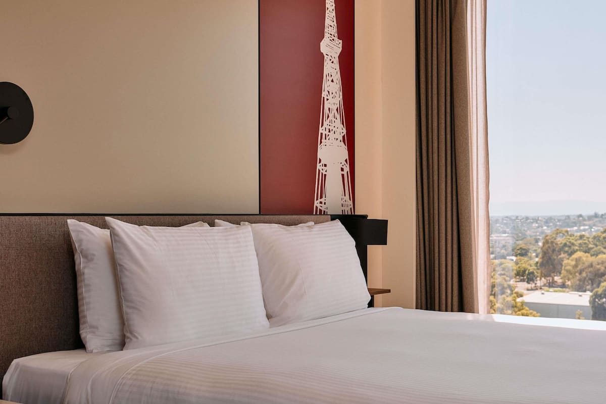 A hotel room with a large bed, a headboard, a nightstand, and a view of a cityscape through the window.