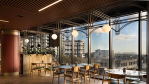 Un interior moderno con suelo de madera, mesas y sillas redondas, amplias ventanas con vista a la ciudad y lámparas colgantes.