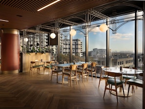 A modern interior with wooden flooring, round tables, and chairs, large windows with a city view, and hanging pendant lights.