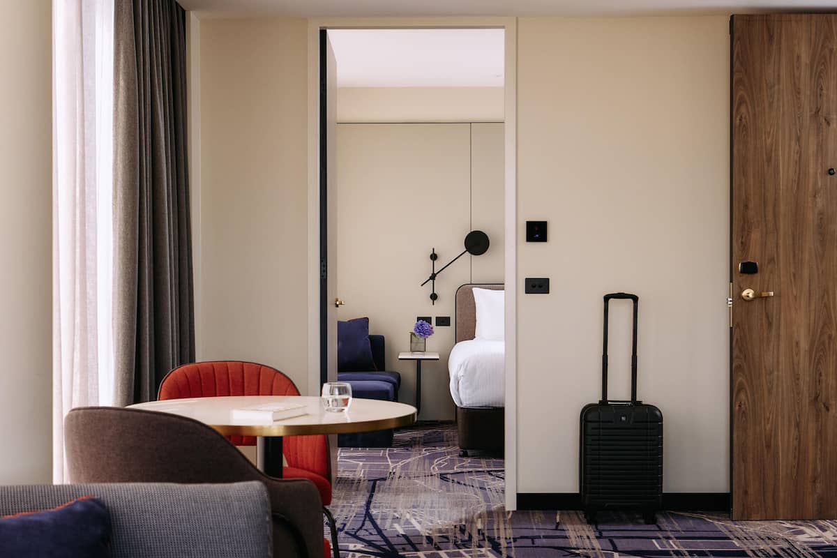 A hotel room with a bed, a desk, a chair, a table, a suitcase, and a window with curtains.