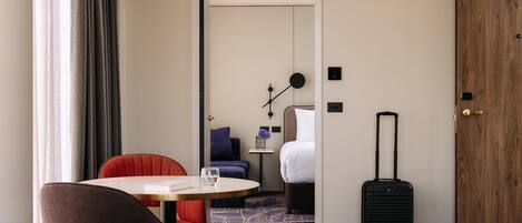 Habitación de hotel con cama, escritorio, silla, mesa, maleta y ventana con cortinas.