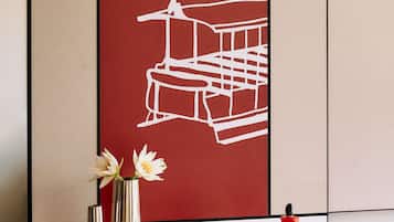 A room with a red wall, a white line drawing of a building, two vases with flowers, and a red bottle on a table.