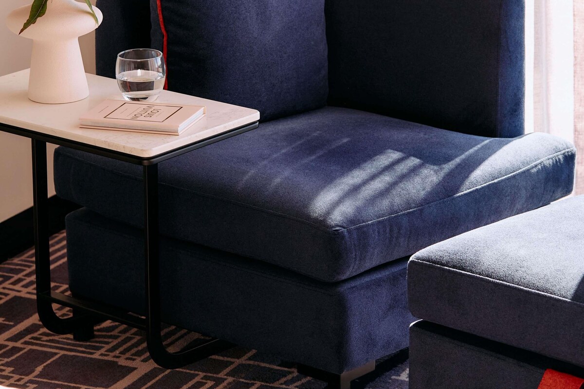 Un sillón azul oscuro con un reposapiés a juego, un jarrón blanco con flores rojas, un vaso de agua y un libro sobre una mesita.