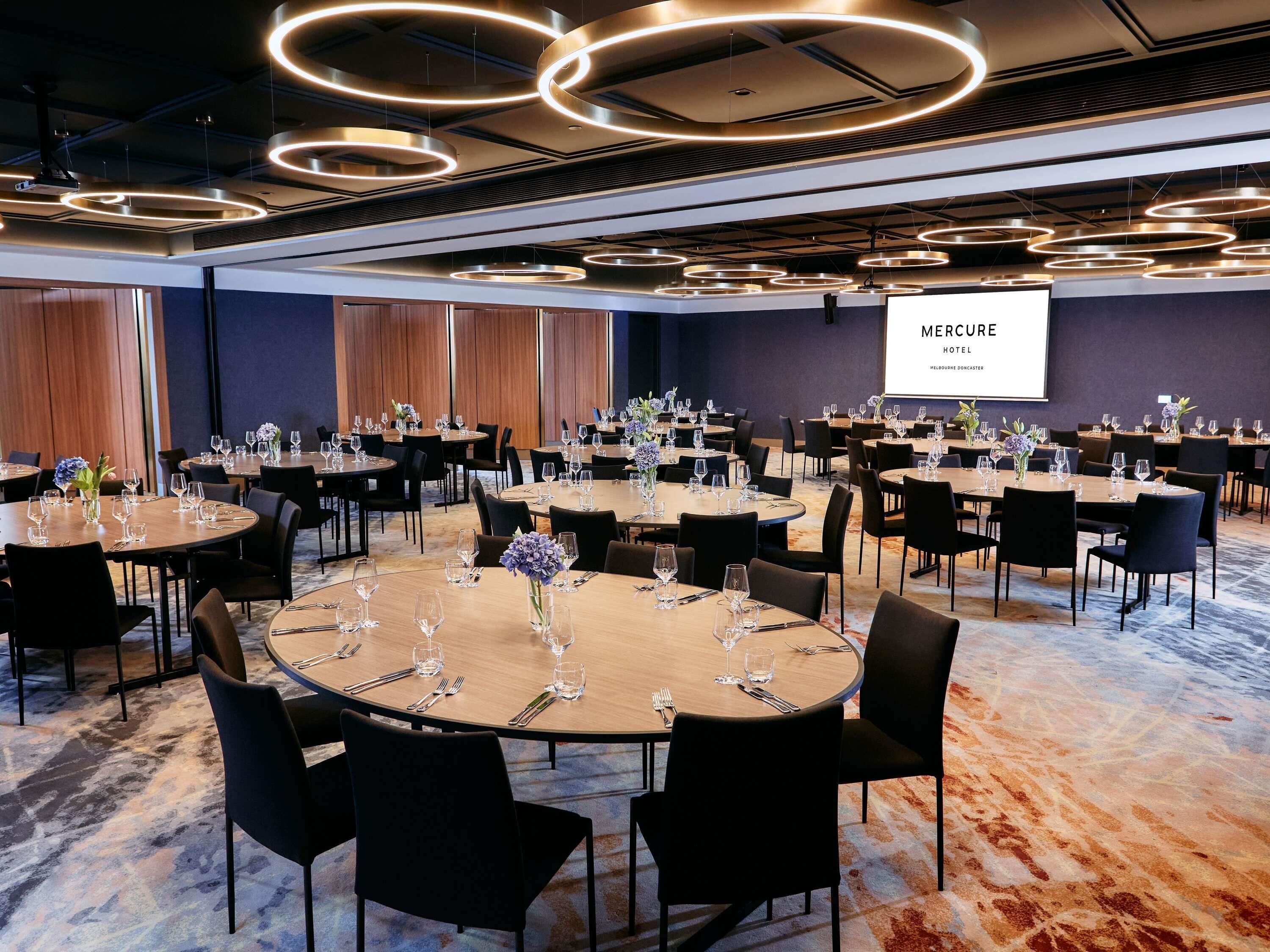 A conference room with round tables, black chairs, and a projector screen displaying 'MERCURY HOTEL.'
