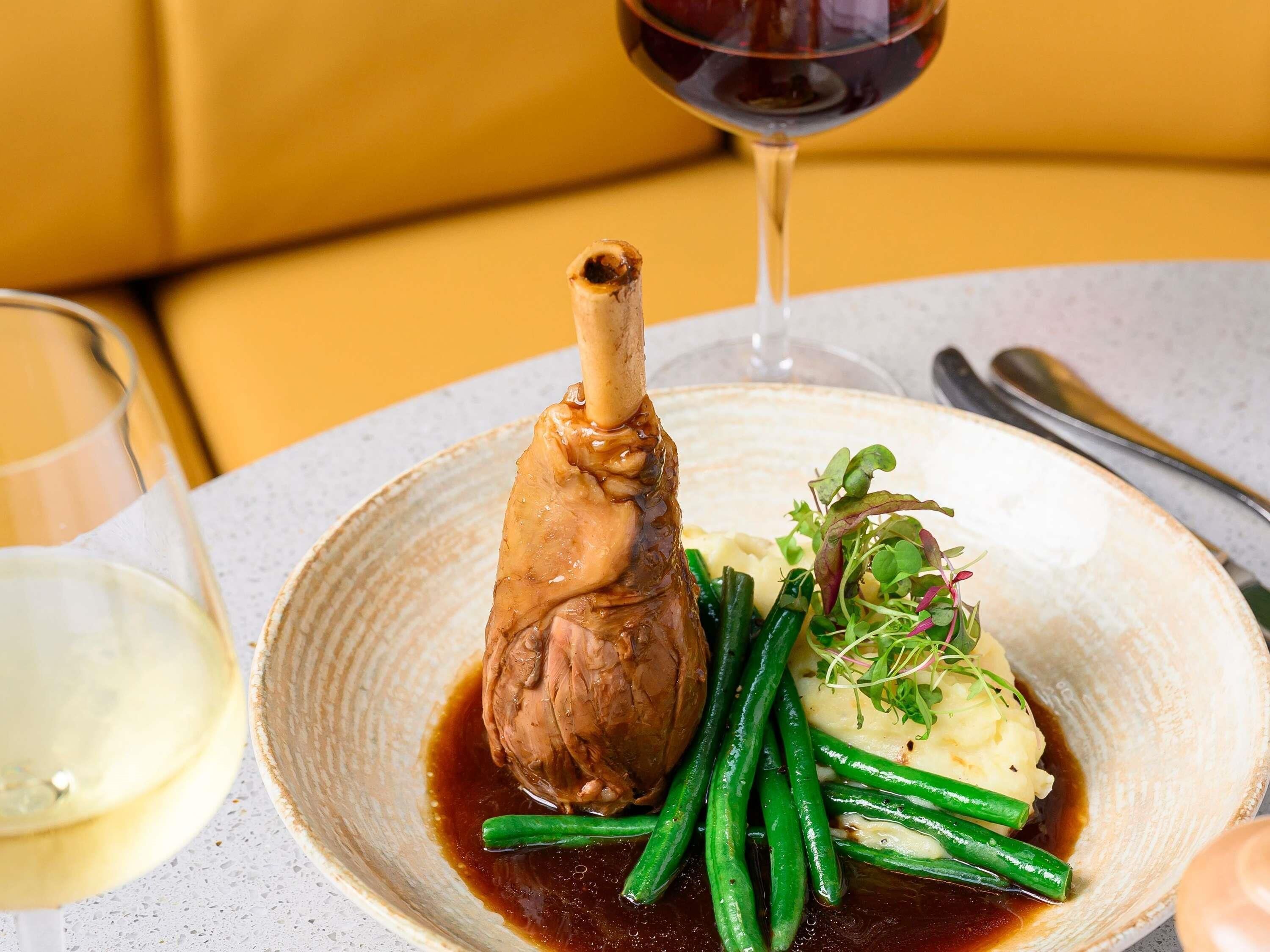 A plate with a bone-in lamb shank, green beans, and mashed potatoes.