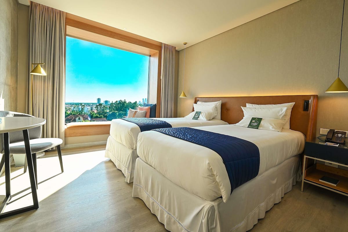 Une chambre d’hôtel avec un grand lit, une petite table et une vue sur le paysage urbain, visible à travers une grande fenêtre.