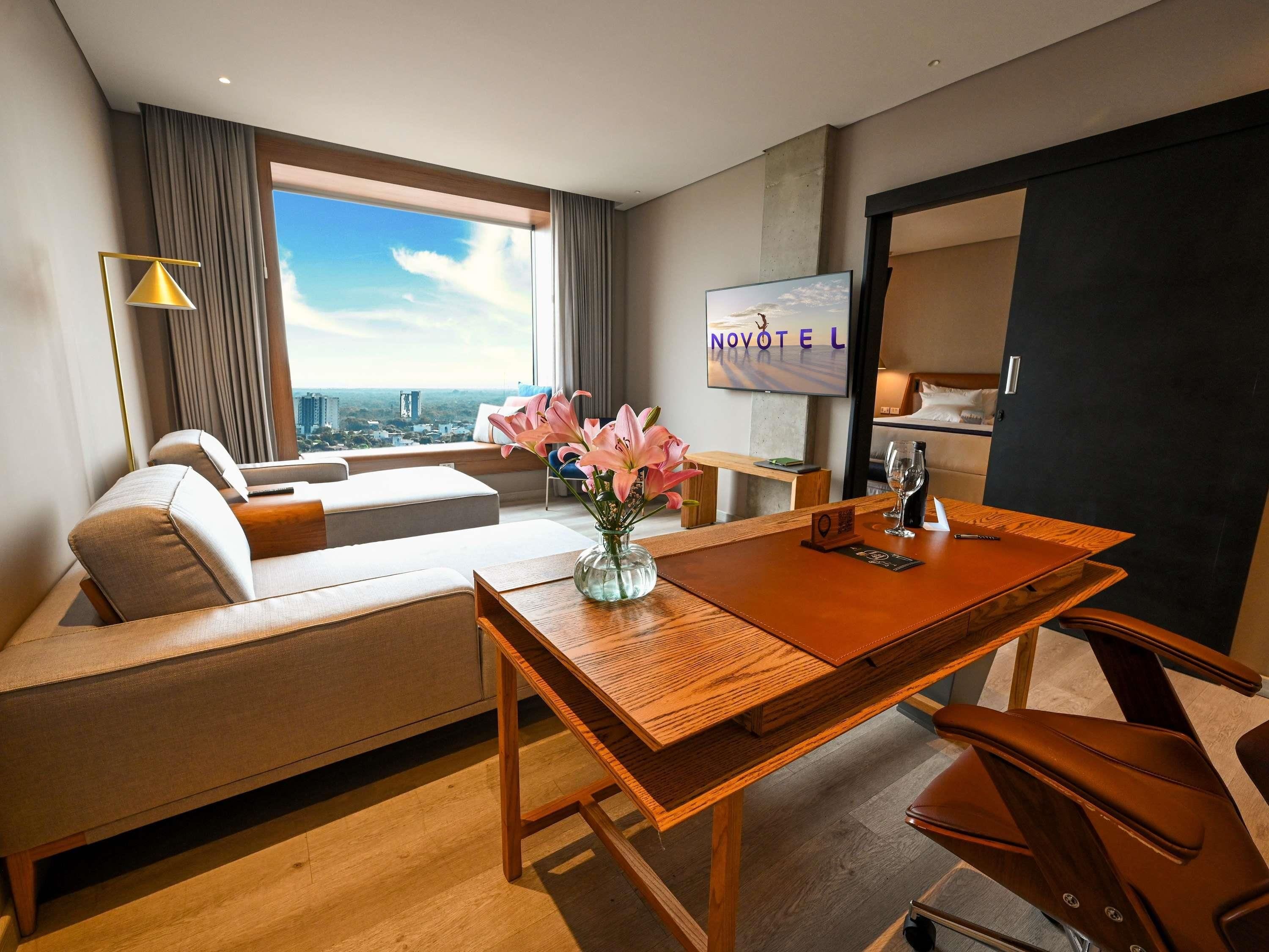 A hotel room with a sofa, a dining table, a TV, and a view of the city.