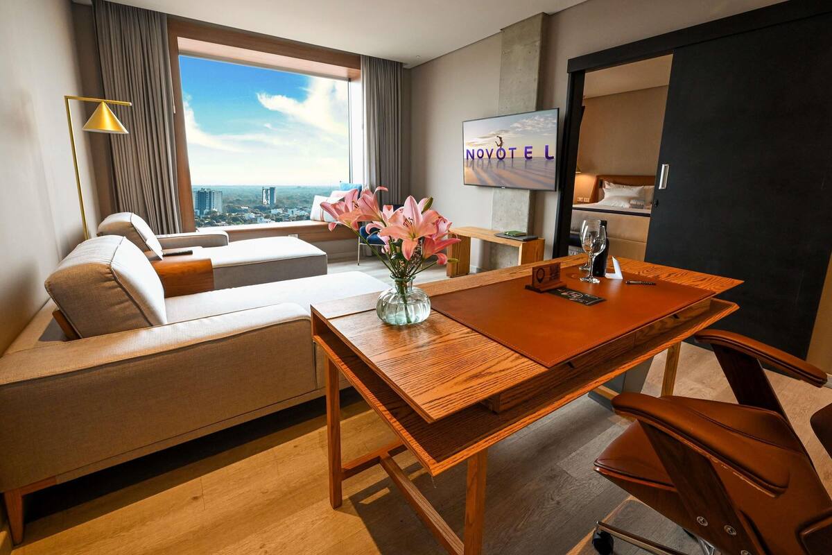 Une chambre d’hôtel moderne dotée d’une grande fenêtre donnant sur la ville, d’un canapé, d’une table à manger agrémentée d’un vase de fleurs et d’un téléviseur fixé au mur.