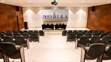 A conference room with rows of chairs facing a stage, a podium, and a large screen displaying 'NOVOTEL'.