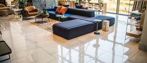 A modern lobby with a large sectional sofa, a coffee table, and a seating area with chairs. There are potted plants and a view of the outdoors through large windows.