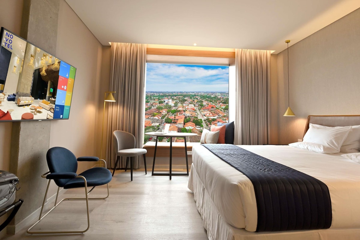 A hotel room with a large bed, a desk, a chair, and a view of a cityscape.
