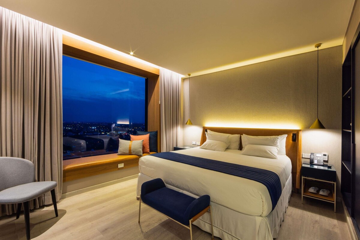 Een hotelkamer met een groot bed, een zithoek, uitzicht op de stad 's nachts en een telefoon op het nachtkastje.
