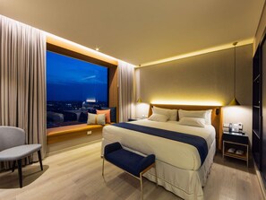 Une chambre d’hôtel avec un grand lit, un coin salon, une vue sur la ville la nuit et un téléphone sur la table de chevet.