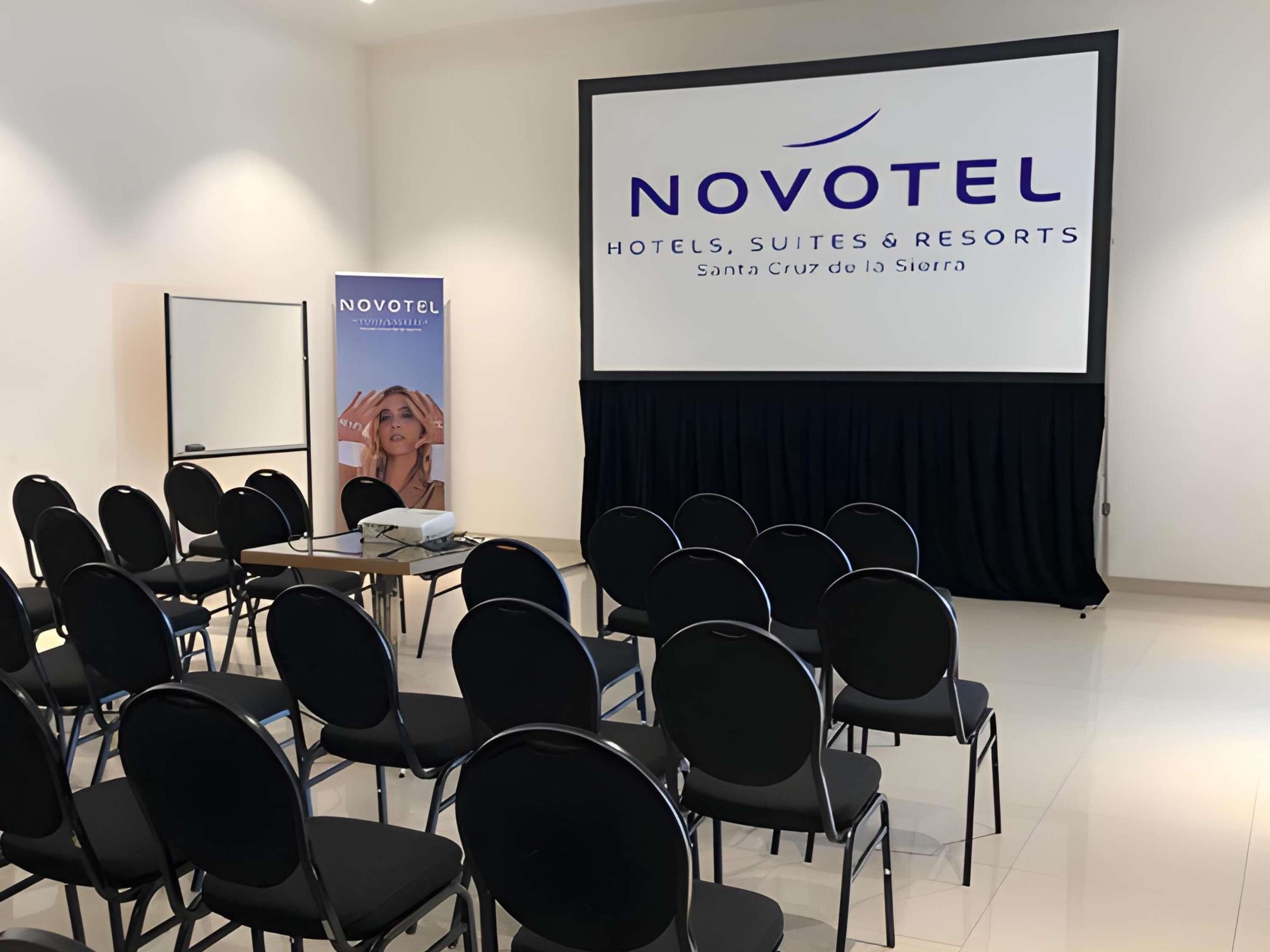 Et mødelokale med sorte stole arrangeret i rækker, et projektorskærm der viser 'NOVOTEL HOTELS, SUITES & RESORTS Santa Cruz de la Sierra', og et banner med NOVOTEL-logoet.