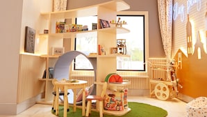 Een goed verlichte kamer met een groot raam, een boekenkast, een speelhoekje met een tafeltje en stoelen, en een radiator.