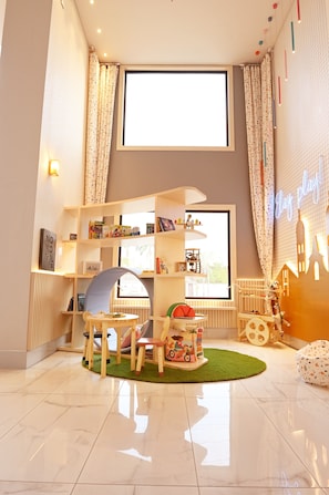Une pièce bien éclairée, dotée d’une grande fenêtre, d’une bibliothèque, d’un espace de jeux avec une petite table et des chaises, et d’un radiateur.