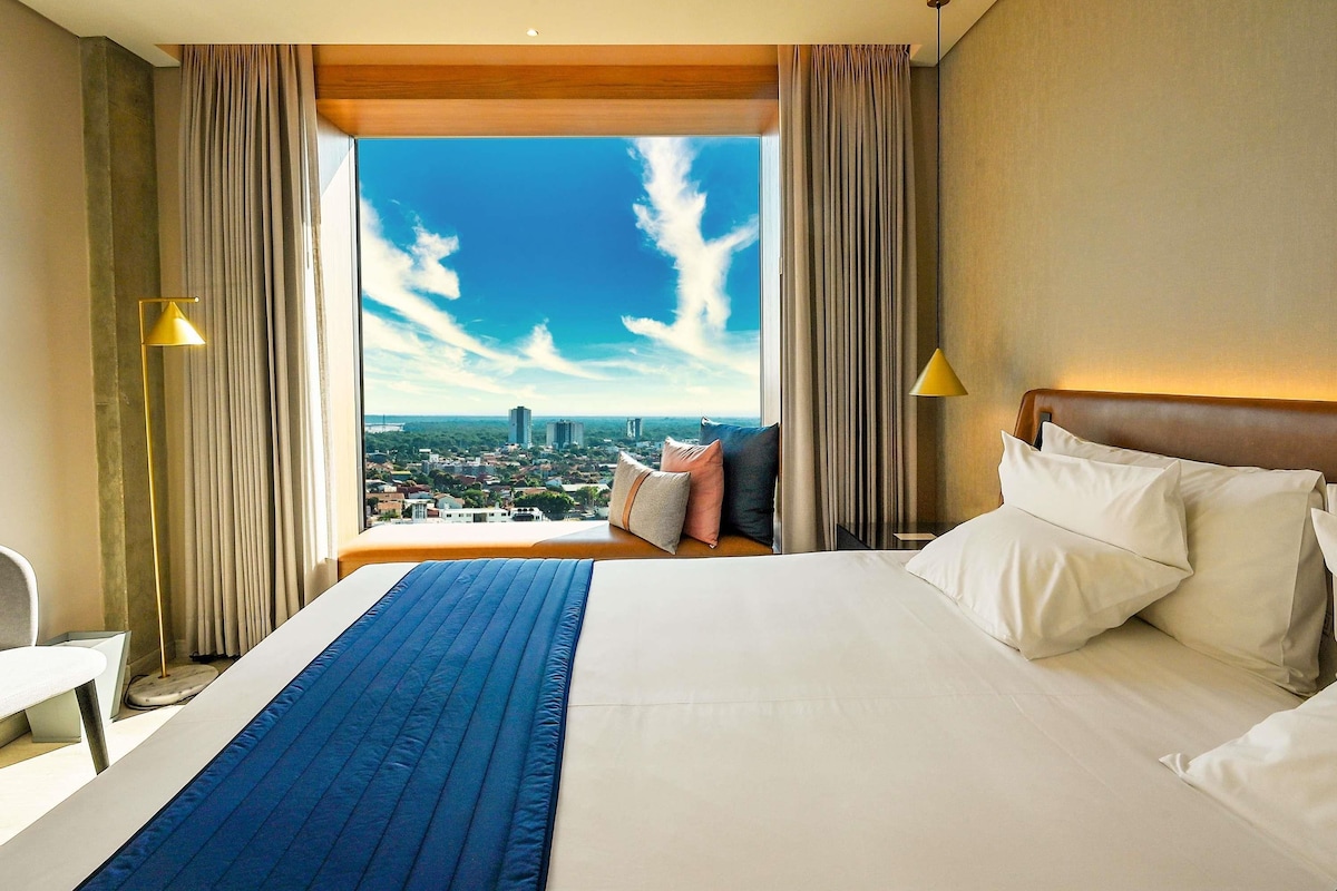 A hotel room with a large bed, a view of the city, and two lamps.