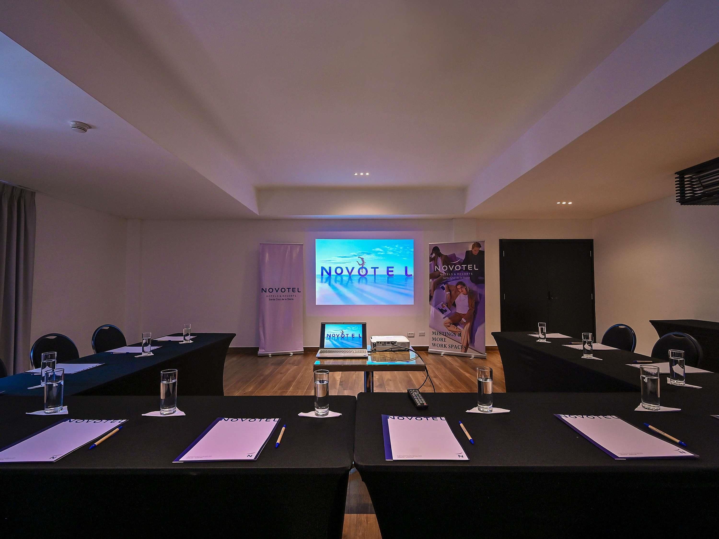 Un salón de conferencias con una pantalla grande que muestra 'NOVOTEL', un cartel con 'NOVOTEL' y 'REUNIONES Y ESPACIOS DE TRABAJO', y un ordenador portátil sobre la mesa.