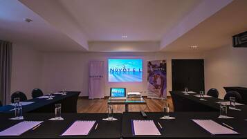 Un salón de conferencias con una pantalla grande que muestra 'NOVOTEL', un cartel con 'NOVOTEL' y 'REUNIONES Y ESPACIOS DE TRABAJO', y un ordenador portátil sobre la mesa.