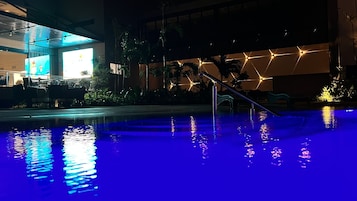 Un área de piscina nocturna con superficie de agua iluminada en azul, palmeras y un edificio moderno con patrones de luz geométricos al fondo.