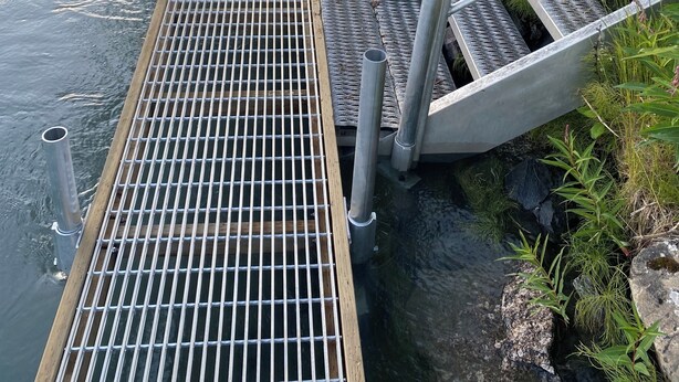 A wooden dock extending into a river, with metal railings and a metal grid floor.
