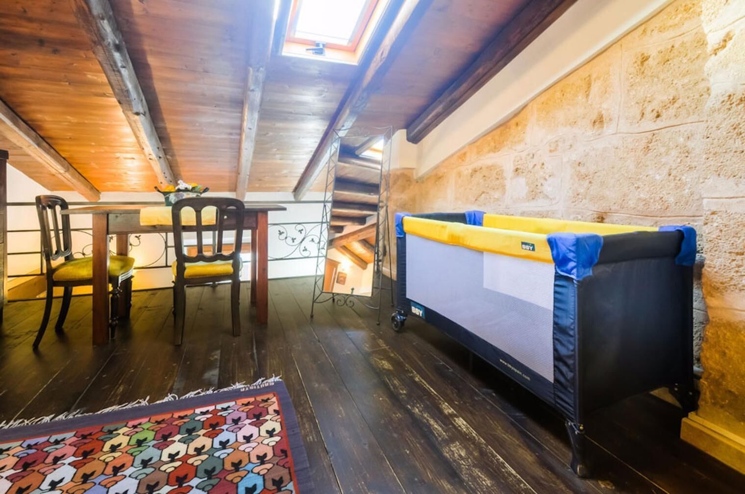 A cozy attic room with a wooden ceiling, a dining table with chairs, a colorful rug, and a large mattress with a canopy.