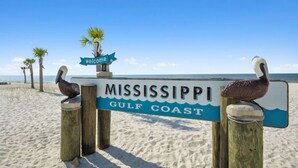 Un letrero de madera con 'MISSISSIPPI GULF COAST' y un cartel de 'welcome', con una playa y palmeras al fondo.