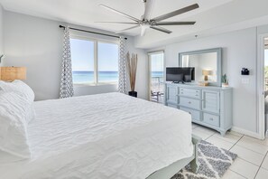 A bedroom with a large bed, a ceiling fan, a TV, a dresser, and a view of the ocean.