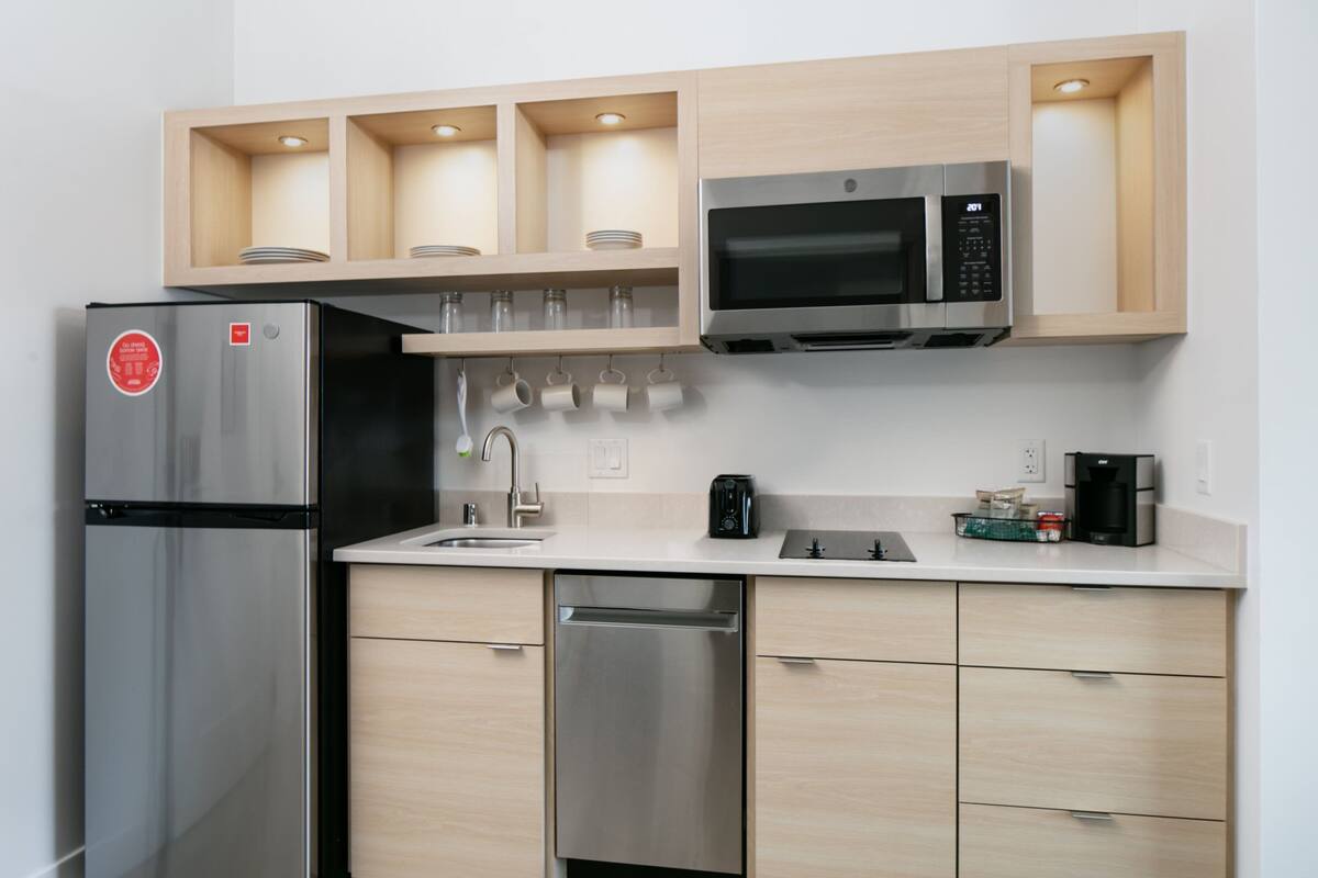 Een moderne keuken met een roestvrijstalen koelkast, magnetron en vaatwasser. Boven het aanrecht zijn houten kastjes en een set hangende mokken.