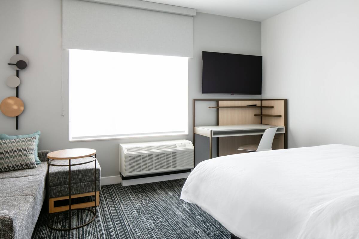 Een moderne hotelkamer met een groot bed, een bureau, een flatscreen-televisie, een raam met jaloezieën, een wandairconditioner en een kleine ronde tafel.