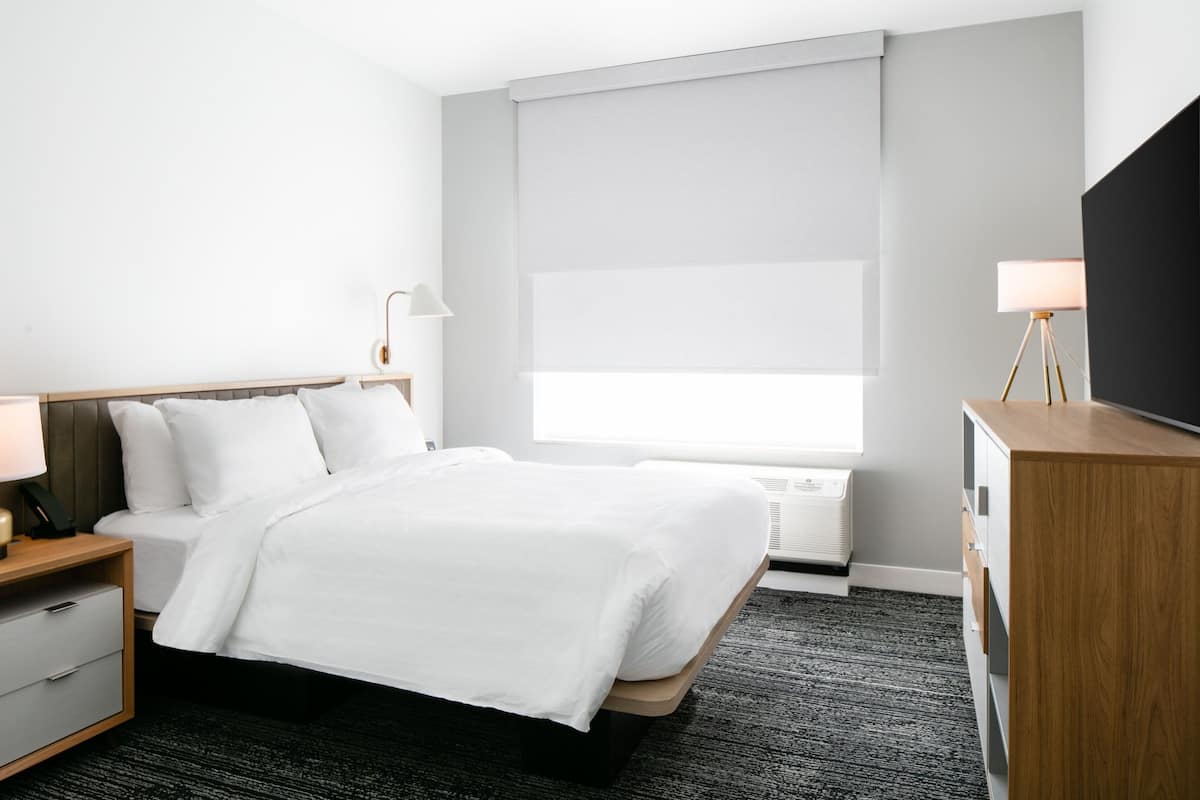 Een hotelkamer met een bed, nachtkastjes, een bureau en een groot raam met zonwering.
