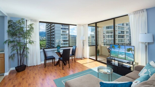 Een moderne woonkamer met balkon, houten vloer, een eethoek en een tv waarop een tropisch strand te zien is.