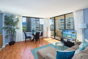 A modern living room with a balcony, wooden flooring, a dining area, and a TV displaying a tropical beach scene. - Charming Waikiki Skytower (Honolulu)