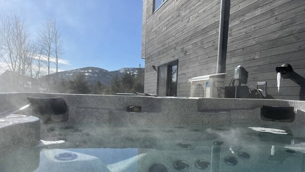 Ett bubbelbad med ånga, en byggnad av trä och en bergig bakgrund.