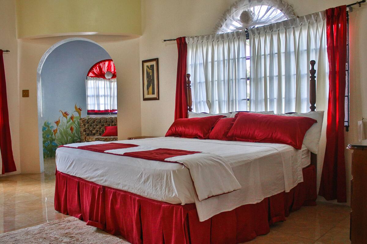 A large bed with red and white bedding, a red curtain, and a window with white curtains.