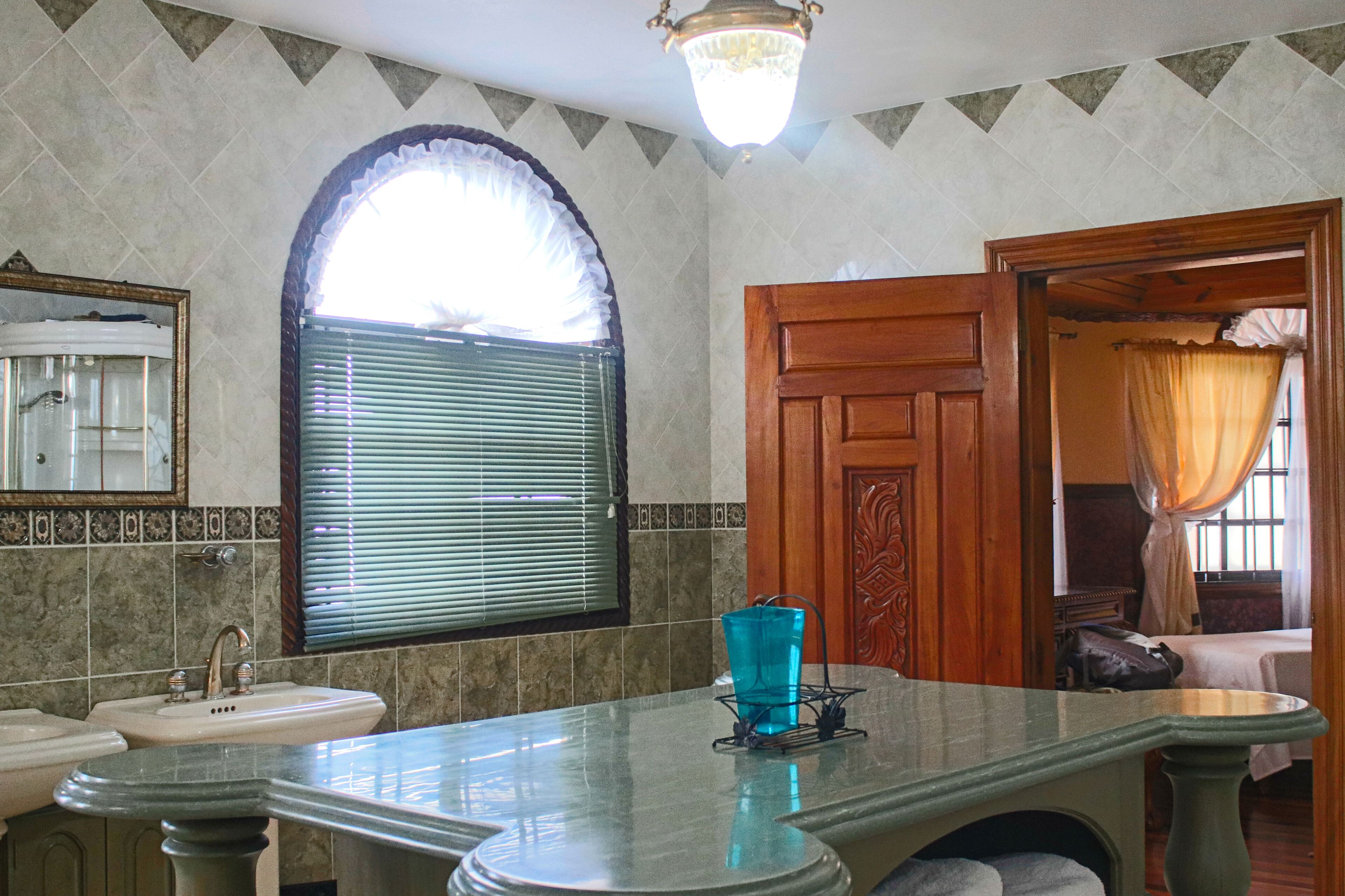 A bathroom with a marble sink, a mirror, a window with blinds, and a wooden door leading to a bedroom.