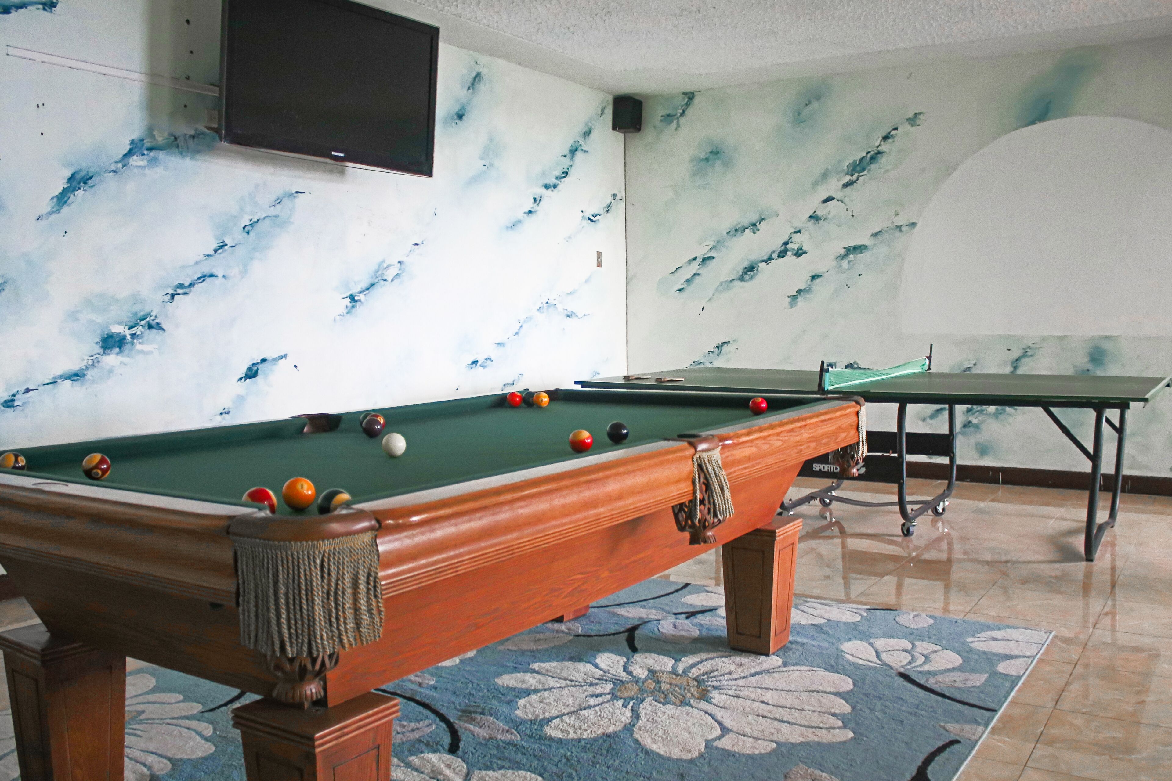 A pool table with balls, a television mounted on the wall, and a marble-patterned wall.