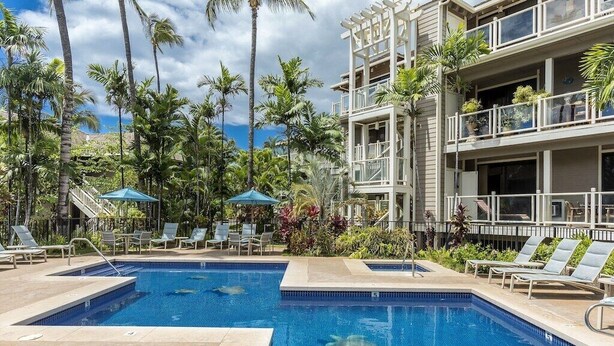 Piscina con sillas de descanso y palmeras, rodeada por un edificio de varios pisos con balcones.