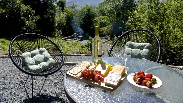 Un coin repas aménagé en plein air, avec des chaises en osier, une table en verre et une variété de mets, notamment du fromage, des raisins et des fraises.