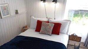 Une chambre avec un lit blanc, des oreillers rouges à motifs, une couverture bleu foncé, une tête de lit blanche, une table de chevet avec une lampe, et une vue sur la verdure à l’extérieur.