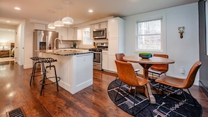 A modern kitchen with a central island, bar stools, a dining table with chairs, and a large window. - The Johnson Chalet in Bristol/Canandaigua*HOT TUB* (Canandaigua)
