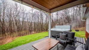 A covered outdoor deck with a hot tub, seating area, and a view of a forested area. - The Johnson Chalet in Bristol/Canandaigua*HOT TUB* (Canandaigua)