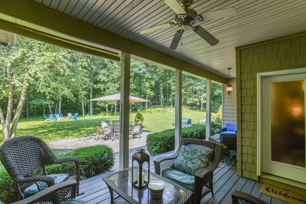 A covered outdoor seating area with wicker furniture, a glass table, and a view of a backyard with a barbecue and patio furniture.