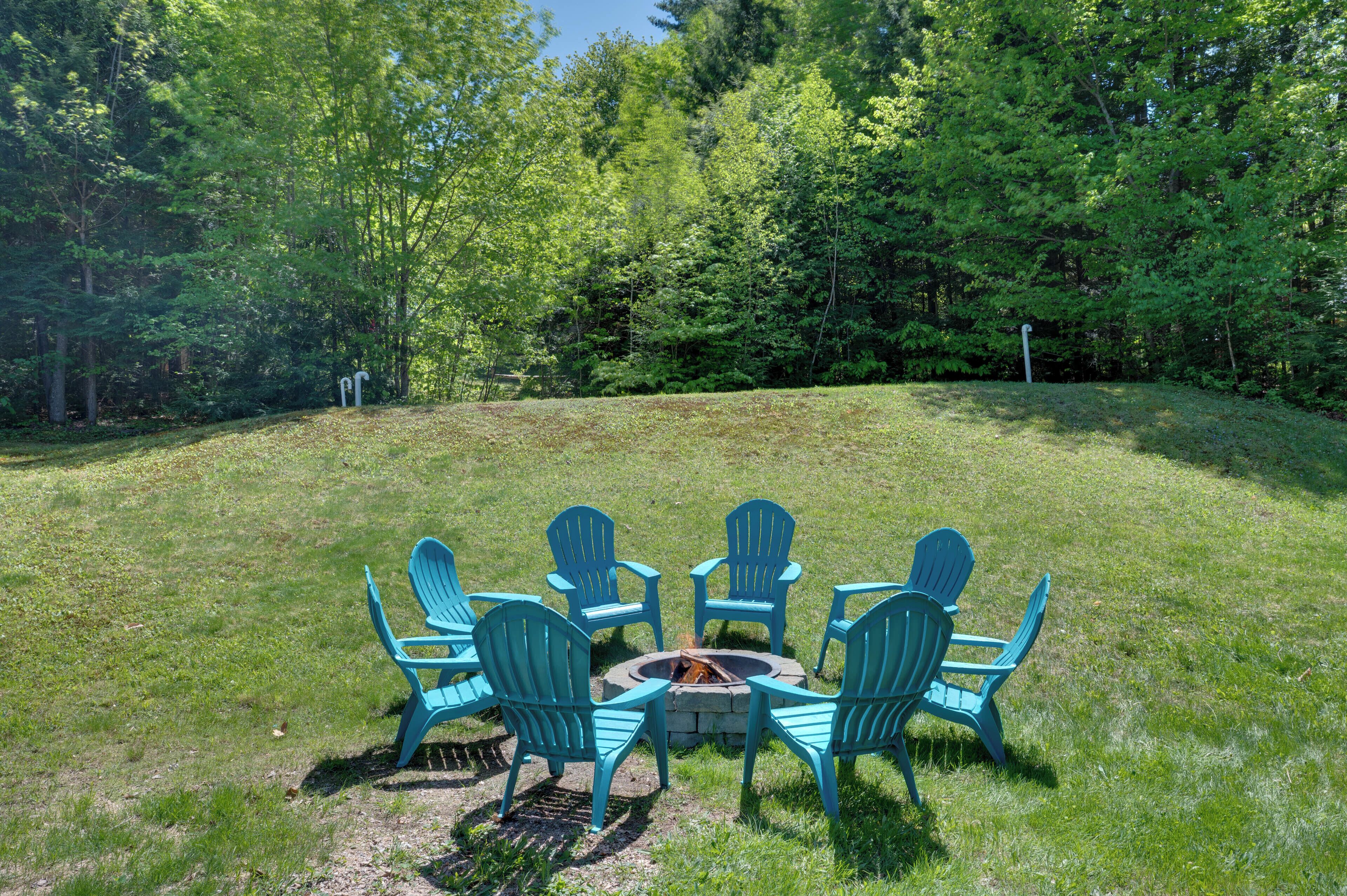 A grassy outdoor area with a circular fire pit and six blue Adirondack chairs arranged around it.