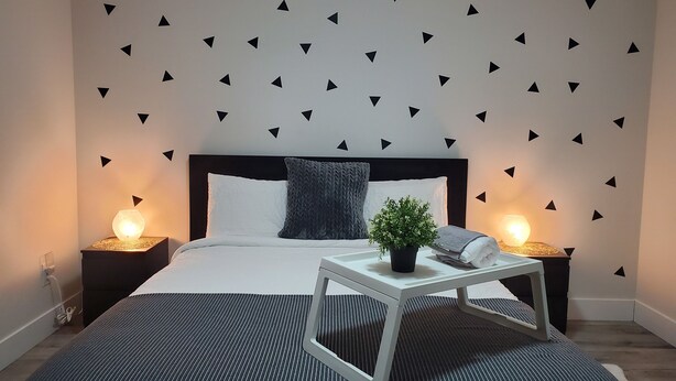 Un dormitorio moderno con una cama, dos mesitas de noche, una lámpara auxiliar y una planta en maceta, además de una pared decorada con triángulos negros.
